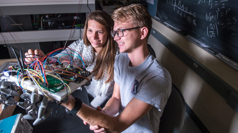 Two engineering students holding up a circuit board and adjusting wires.
