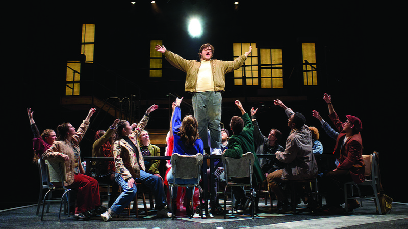 A scene from the Rent performance. A man is standing on a table surrounded by many people sitting in chairs.