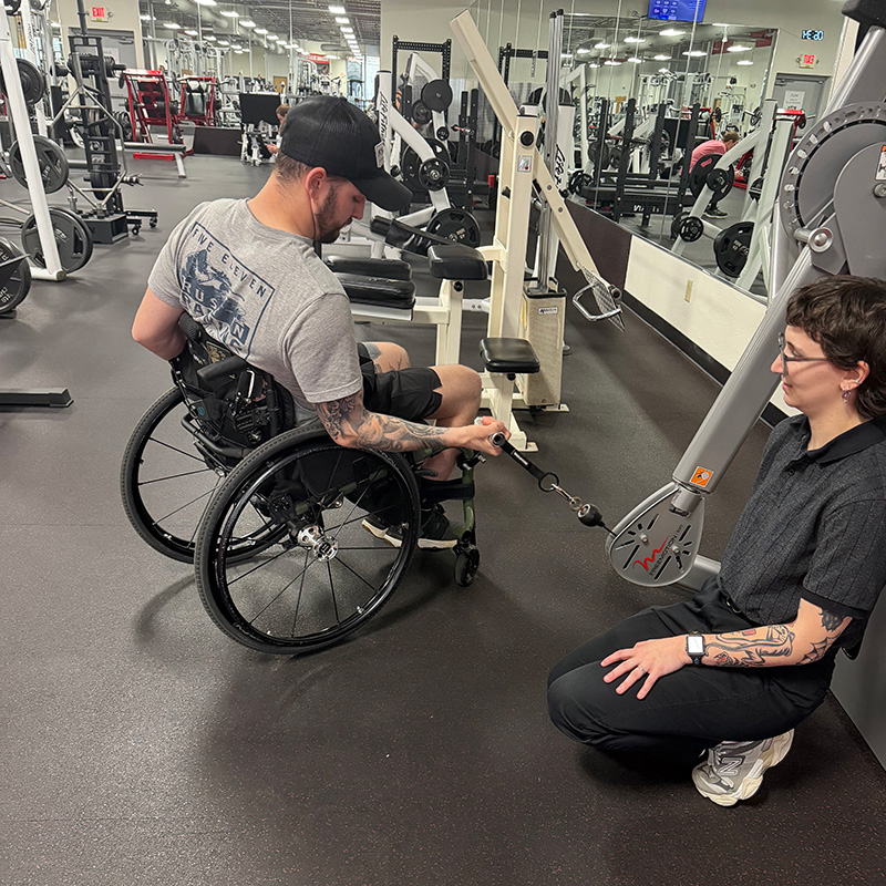 Spinal cord injury patient in wheelchair uses a strength resistant machine with guidance from UE Physical Therapy student.